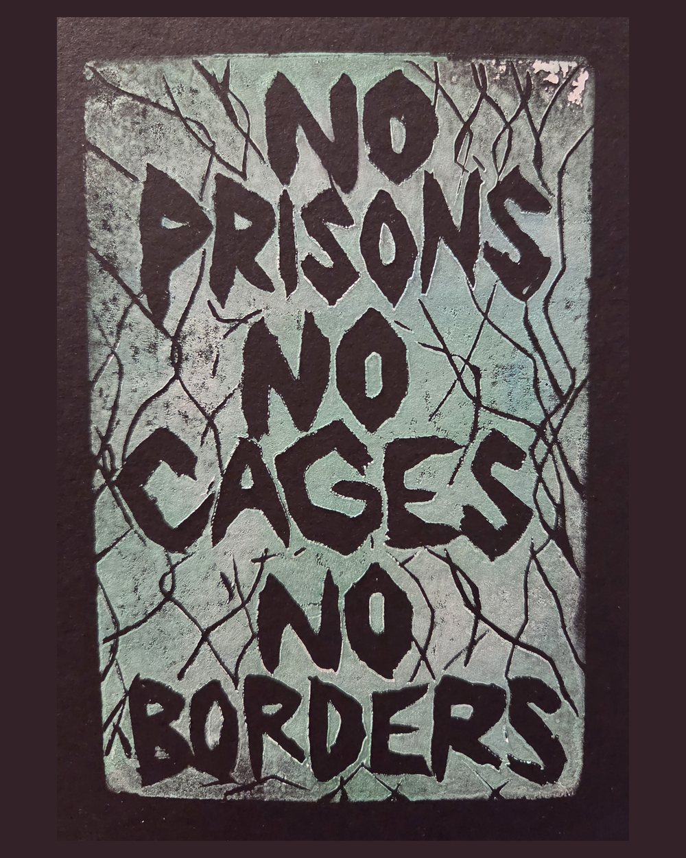 Linocut printed in pale teal on black paper: four lines of hand-carved capitals reading NO PRISONS NO CAGES NO BORDERS, behind a broken chain-link fence.