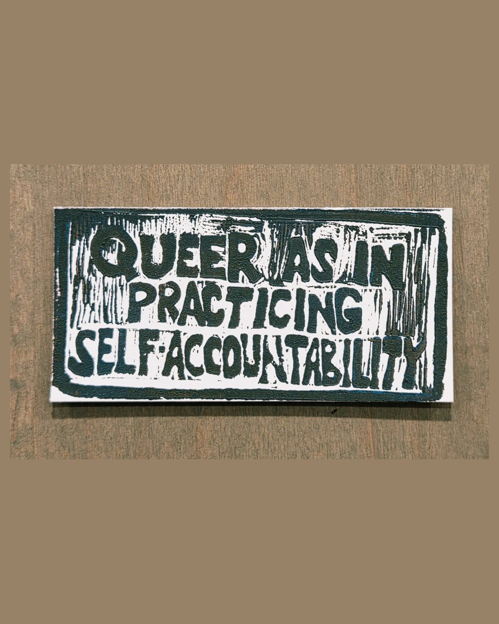 Block print in dark teal on white: three lines of hand-carved capitals reading QUEER AS IN PRACTICING SELF-ACCOUNTABILITY, inside a heavy carved border.