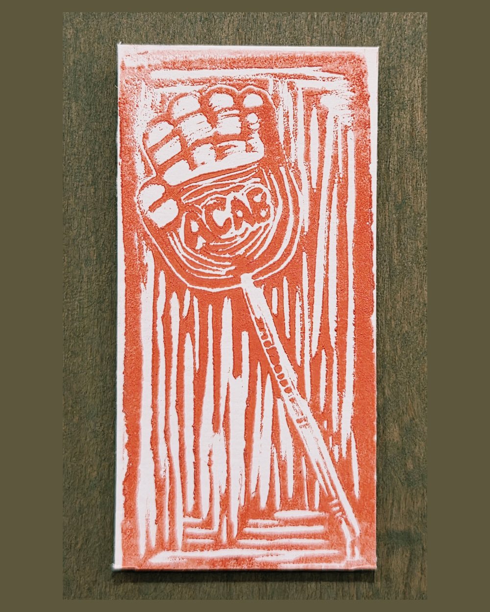 Linocut in red-orange: a raised fist gripping a staff, with ACAB carved into a circle at its centre, surrounded by vertical gouge marks.