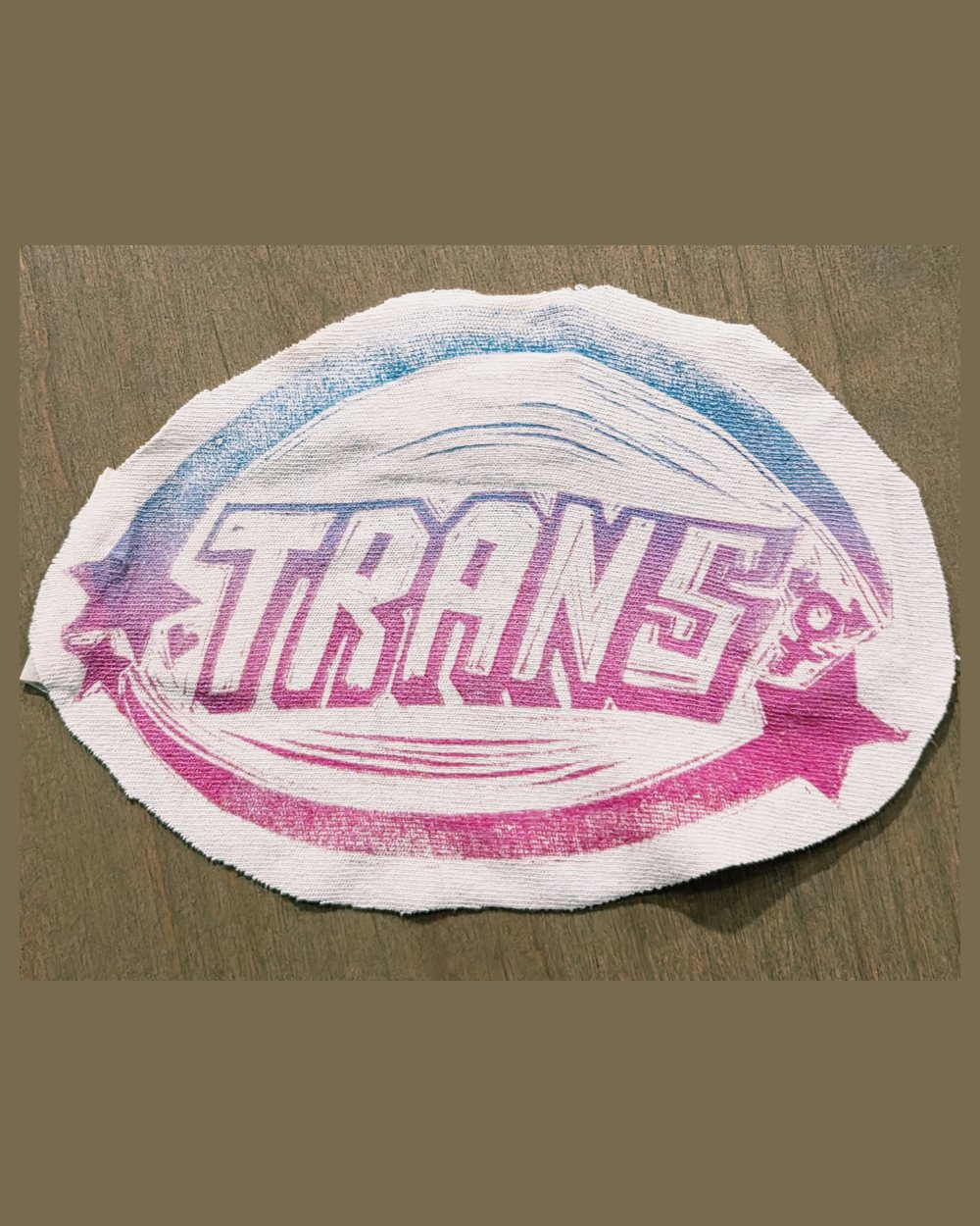 Block print on canvas, cut to an oval patch: the word TRANS in carved capitals inside a ring, printed in a pink-to-blue blend with stars at each end.
