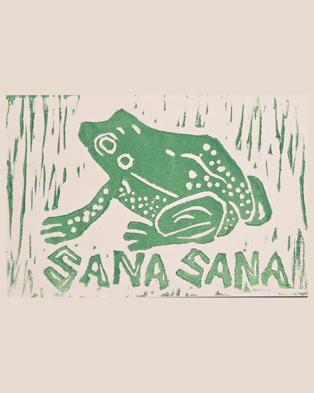 Linocut of a spotted frog printed in green ink on cream paper, with SANA SANA lettered below and gouge marks like reeds around it.